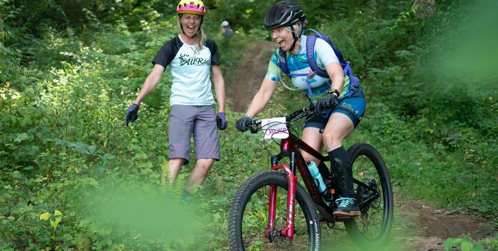 Two women laughing, one riding while the other stands nearby to coach her