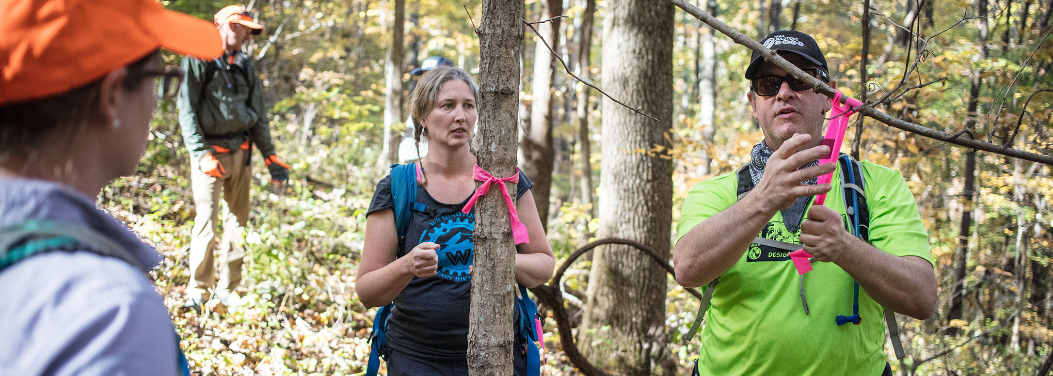 One planner in green shirt ties a flagline to a tree to indicate a future trail path as two planners look on