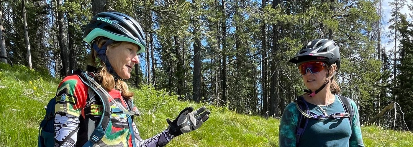 Women of Sand Point, Idaho in mountain biking gear talking trails in a green grass field surrounded by pines. 