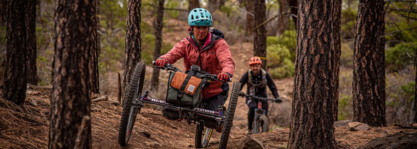 A brunette woman adaptive mountain biker weaves through trees and is followed by a male mountain biker.