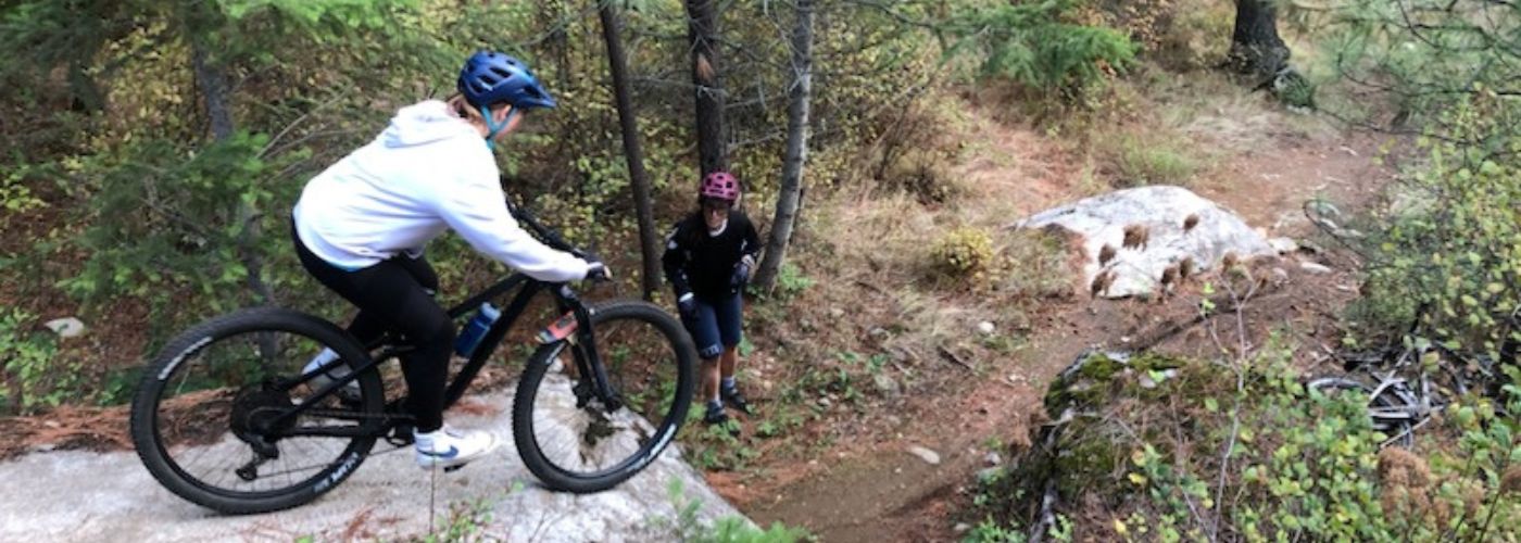 Lady mountain biker contemplating a medium rock drop while another rider monitors.