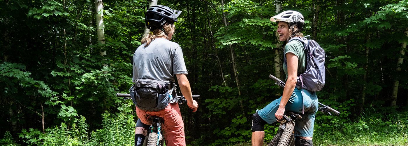 Two women at a trailhead talking and laughing on their bikes. They are muddy and happy.