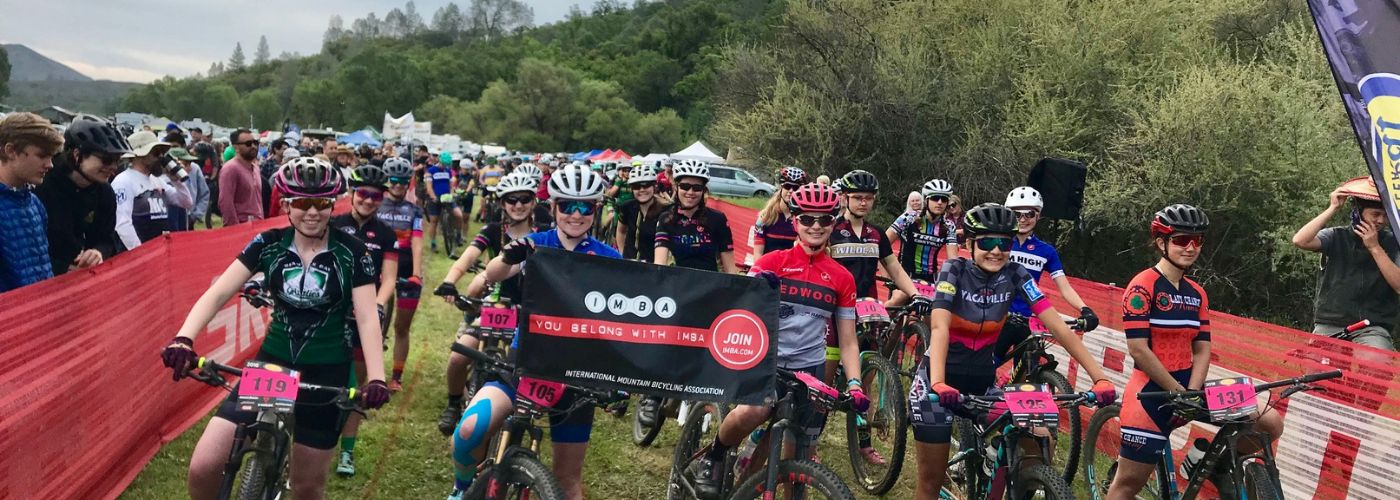 start of women's mountain bike race with pink race numbers and a host of women racers holding IMBA banner.