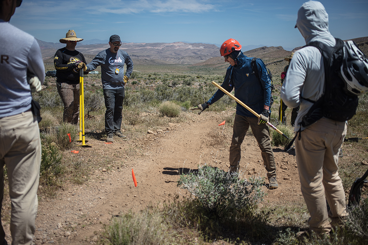 Six people surrounding a trail with tools in hand, participating in an IMBA Trail Care Workshop in 2023.