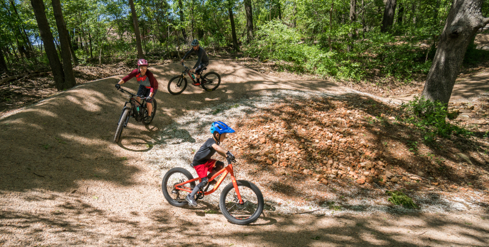 a group of three with a young kid in the lead rides their bikes around a turn with inslope