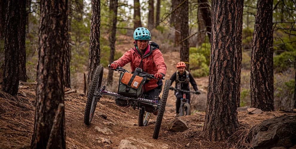 A brunette woman adaptive mountain biker weaves through trees and is followed by a male mountain biker.