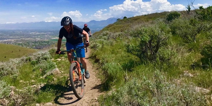 Mountain biker with blue shorts rides along bench cut trail, overlooking a Salt Lake Valley community