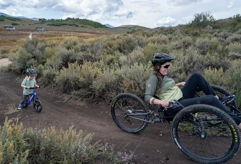 Amanda on an adaptive mountain bike followed by her daughter Zoey on a Strider bike, riding on a dirt trail