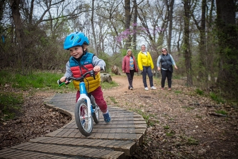 Young kid riding over a wooden feature on a balance bike smiling while family looks forward smiling in the distance.