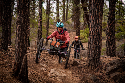 A brunette woman adaptive mountain biker weaves through trees and is followed by a male mountain biker.