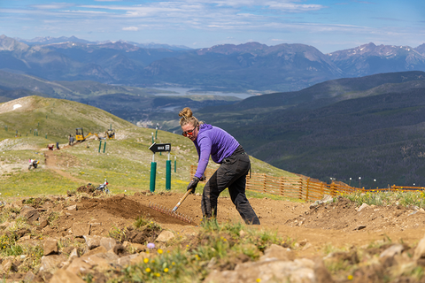 Jess Didion rakes a berm in the foreground as a mini excavator works in the background.