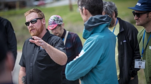 Man wearing sunglasses and a black button down shirt addresses a group of people at a trail education event