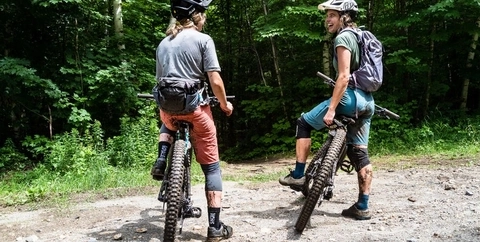 Two women smiling and talking after some muddy mountain biking at a trailhead.