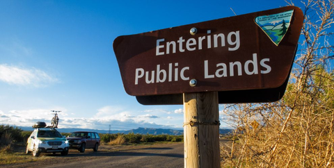 “Entering Public Lands” sign in the forefront of a open vista with a mountain bike on top of a car in the background.|