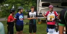 Three youth mountain bikers standing in circle, listening to one young white male mountain biker speak.