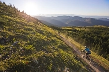 Looking down from above and behind, you see two mountain bikers cruising along the side of a mountain on singletrack. Green hillsides, pine forests, and green mountains are in the distance with a hazy blue sky.