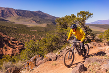Rider in yellow jacket pedals an eMTB in a red dirt, high desert landscape with mountain range in background 