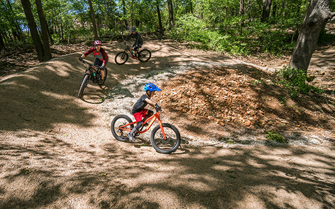 Two parents and a kid mountain biker ride through a berm.