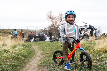 Youth balance bike rider, smiling and standing next to trail. 