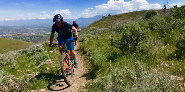 Mountain biker with blue shorts rides along bench cut trail, overlooking a Salt Lake Valley community